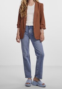 Blazer marron aux manches retroussées, chemise blanche ajustée, jean bleu taille haute, et sandales compensées bleu clair. Tissu lisse et coupe décontractée.