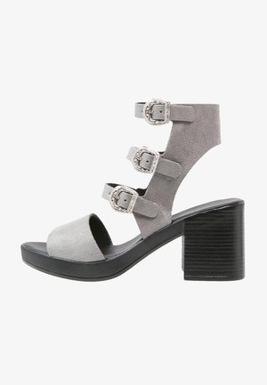 Sandalía de tacón alto de ante gris con tres hebillas plateadas ajustables y un tacón grueso negro.