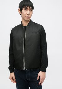 Crna bomber jakna sa zatvaračem sprijeda, rebrastim manžetnama i izvezenim logotipom na prsima. Nošena preko crne majice s dugim rukavima.