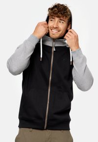 Sudadera con cremallera en negro y gris, con un bolsillo tipo canguro, mangas en contraste y cordones blancos. Presenta una textura de tela suave.