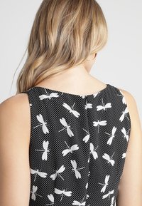 Svart ärmlös klänning med tryck av vit trollslända på en prickig bakgrund. Mjuk tyg, knappdetalj på ryggen och rundad halsringning.