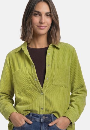 Mujer con el cabello castaño hasta los hombros, que lleva una camisa de pana verde lima sobre una camiseta oscura y vaqueros azules con las manos en los bolsillos.
