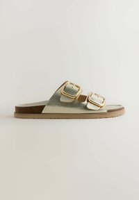 Sandale plate à semelle glissante avec deux brides métalliques dorées à boucle et une semelle marron coussinée, présentée en profil latéral sur un fond neutre.