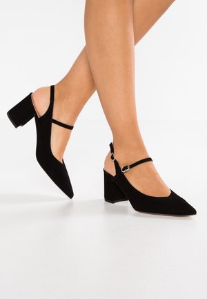 Vrouw draagt zwarte suède schoenen met blokhak en enkelbandjes, staat met één voet iets omhoog op een witte vloer.
