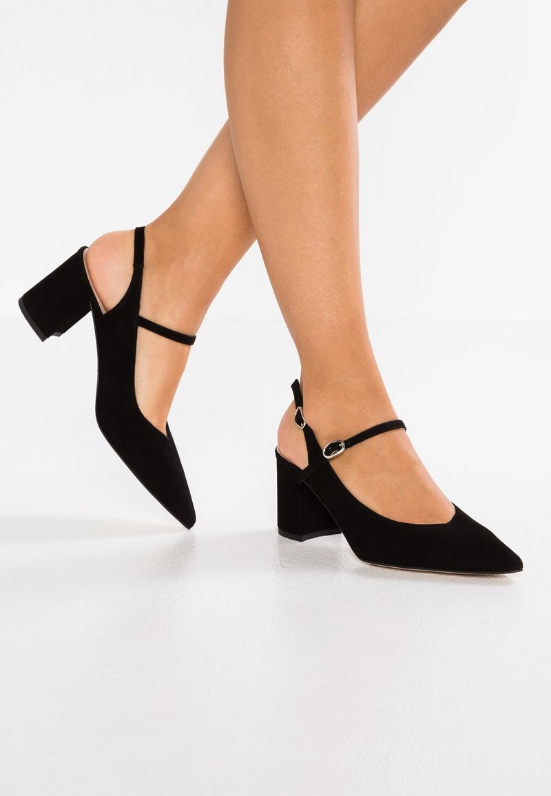 Femme portant des chaussures en suédine noire à talons carrés avec brides à la cheville, debout avec un pied légèrement levé sur un sol blanc.