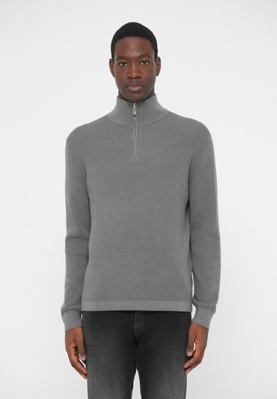 Suéter gris con cremallera y alto cuello, tejido de punto texturizado, puños y dobladillo acanalados, llevado sobre jeans de color gris oscuro, fondo liso.