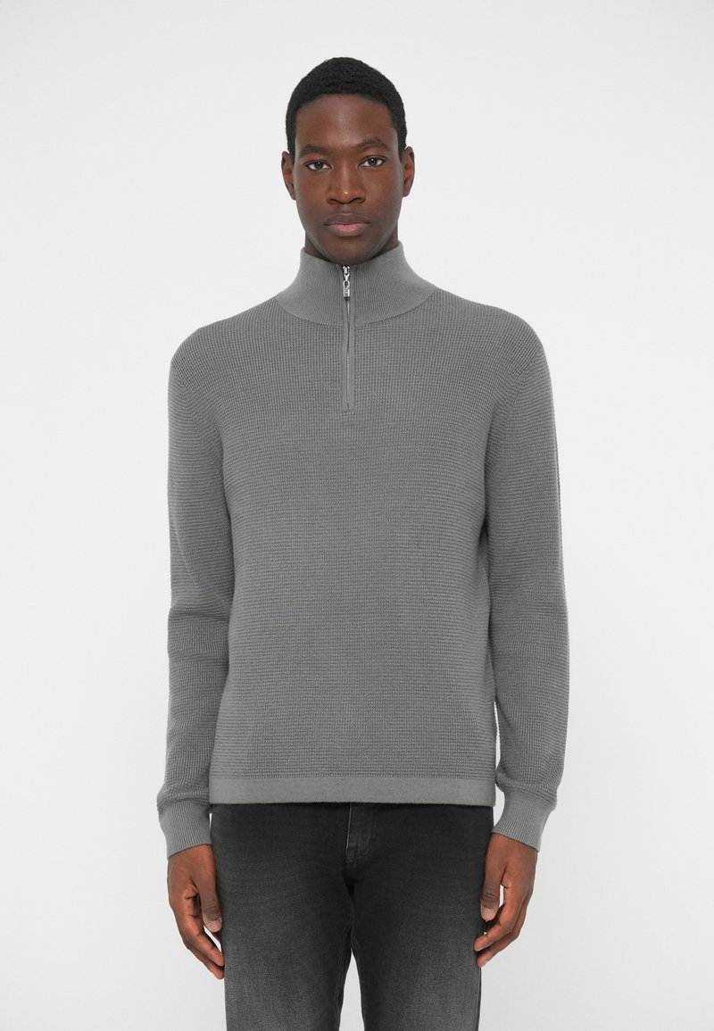 Gray zip-up sweater with a high collar, textured knit fabric, ribbed cuffs and hem, worn over dark gray jeans, plain background.