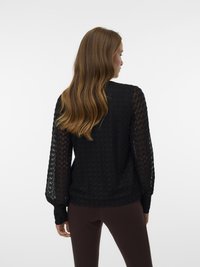 Γυναίκα με μακριά κυματιστά μαλλιά στραμμένη προς τα πίσω, φορώντας ένα μαύρο υφασμάτινο μπλουζάκι μακρύ μανίκι και σκούρες καφέ εφαρμοστές παντελόνες, σε ουδέτερο φόντο.