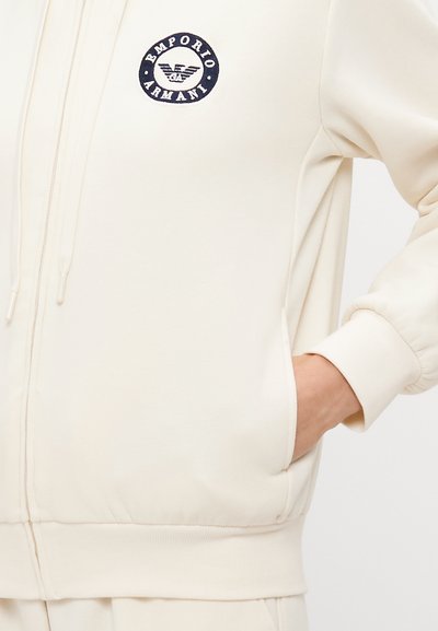 Svijetlosmeđa zip-up dukserica s rebrastim manžetama i donjim rubom. Ima izvezeni mornarski plavi okrugli logotip na lijevoj strani prsiju. Uključuje bočnu džep.