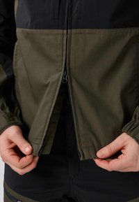 Mains tirant la fermeture éclair d'une veste bicolore noire et vert olive avec des poignets élastiques et un pantalon noir visible en dessous.