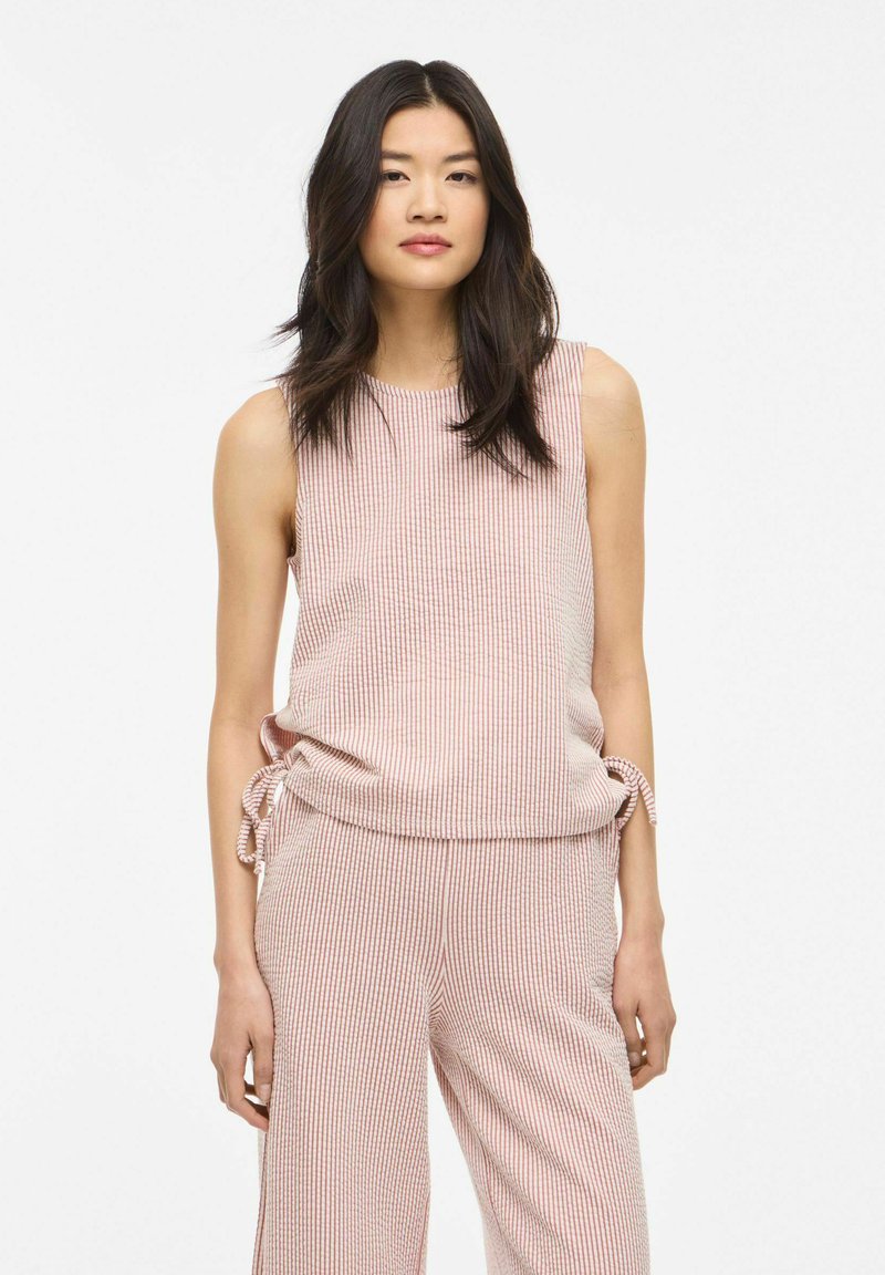 Noor naine pikkade tumedate juustega, kes kannab roosa-valget varrukateta ülaosa ja sobivaid laia säärega pükse küljetillukestega.