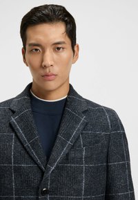 Blazer a quadri grigio scuro con tessuto testurizzato e revers a notched, caratterizzato da un singolo bottone nero e un dolcevita blu navy a contrasto sotto.