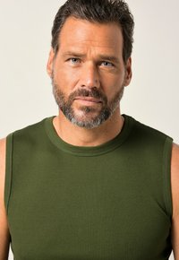 Canotta a coste senza maniche di un verde scuro, con scollatura rotonda e texture liscia, indossata da un uomo con barba e capelli corti.