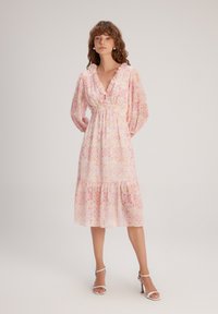 Robe à imprimé floral en rose clair avec un col froncé, taille élastique, manches bouffantes longues et ourlet à volants. Portée avec des sandales à talons blanches.