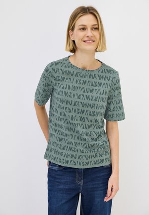 Groene korte mouwen T-shirt met een gestructureerd, abstract patroon in donkerder groen. Ronde halslijn, gecombineerd met blauwe spijkerbroek.
