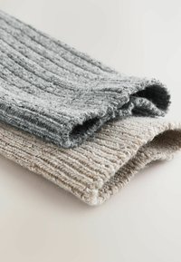 Zwei gerippte Strickstoffmuster, eines grau und eines beige, mit einer weichen Textur und gerollten Kanten, die auf einem neutralen Hintergrund liegen.
