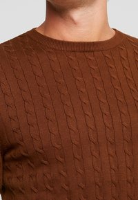 Pull marron à torsades avec un col rond et un motif texturé. Le tissu semble doux et ajusté, mettant en valeur la silhouette.