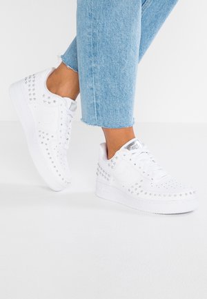 Personne portant un jean court bleu clair et des baskets blanches décorées de clous étoiles argentés sur fond blanc.