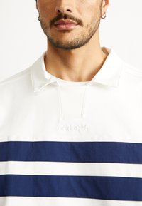 Balts polo krekls ar tumši ziliem horizontāliem svītrām, apkakles kakla daļa un izspiedis Adidas logo uz krūtīm. Texturēts kokvilnas materiāls.