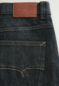 Sötétkék farmernadrág bőr címkével, amely dombornyomott szöveget tartalmaz. Kiemelkedő varrásminták és klasszikus ötlovas zsebes kialakítás.