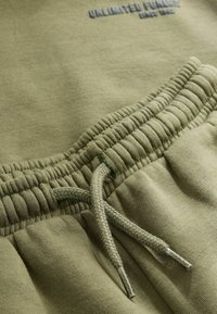 Közelről nézve, olajzöld elasztikus derékpánt húzózsinórral a melegítőnadrágon, bemutatva a textil textúráját és a varrási részleteket.
