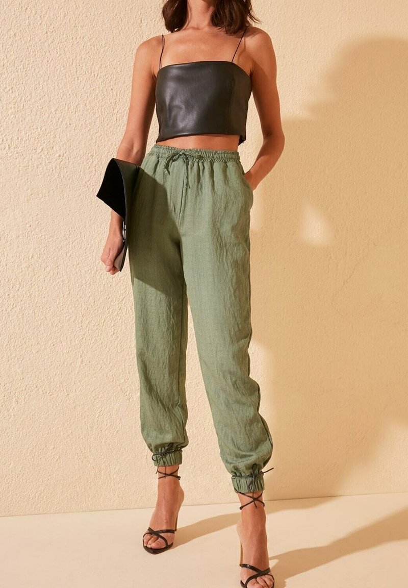 Haut court en cuir noir, pantalon ample vert clair avec poignets élastiques, et talons hauts noirs à lanières. Pochette simple tenue sous le bras.