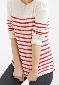 Weißer Strickpullover mit roten horizontalen Streifen, gerippten Bündchen und rundem Halsausschnitt. Kombiniert mit dunklen Jeans.