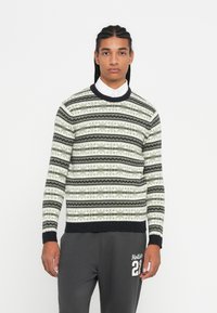 Jonge man met gevlochten haar, gekleed in een gebreide trui met een patroon over een wit shirt en donkere casual broek, met de tekst "Hollister 22".