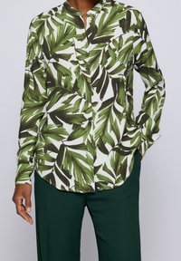 Chemise à manches longues en tissu blanc avec un motif de feuilles tropicales vertes. Dispose d'un col sans col et d'un ourlet incurvé. Assortie à un pantalon vert.