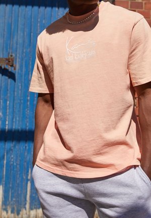 Mann trägt ein hellpfirsichfarbenes "karl kani-jeans" T-Shirt und hellgraue Jogginghose, steht an einer blauen Holztür im Sonnenlicht.