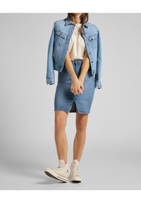 Veste en jean de couleur bleu clair, col pointu et fermeture à boutons, associée à une jupe en jean bleue avec une fente à l'avant, et des baskets montantes beiges.
