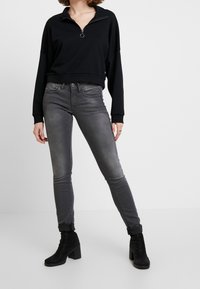 Sudadera negra con cremallera en el cuello, combinada con jeans ajustados grises, que presentan una textura suave y un leve desgaste. Las botas son negras con un tacón ancho.