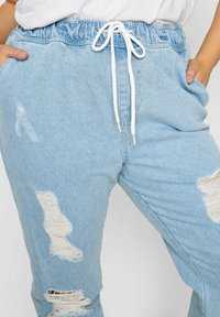 Joggers de mezclilla azul claro con cintura ajustable, que presentan múltiples parches desgastados y bolsillos laterales. Textura suave, corte relajado.