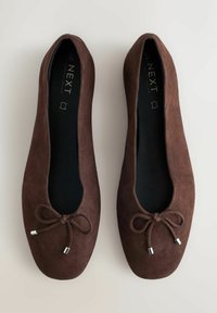 Barna szarvasbőr balettcipő kerek orral, hullámos szegéllyel és az elején díszítő csomóval, amely fémes díszítéseket tartalmaz.