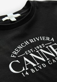 Mustapaita, jossa valkoista kirjontaa: "RANSKAN RIVIERA PER. 1982 CANNES 14 BLVD CAR." Pyöreä kaula-aukko ja pehmeä materiaali.