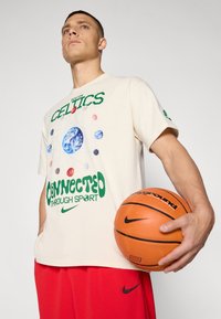 Kremfarget t-skjorte med "BOSTON CELTICS" i grønt, med planeter som grafikk og teksten "CONNECTED THROUGH SPORT" i grønt. Holder en basketball.
