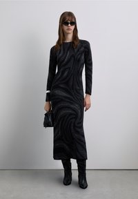 Abito nero a maniche lunghe con motivo a spirale, abbinato a stivali neri al ginocchio e una borsa nera. Il design presenta riflessi scintillanti.