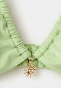 Tissu vert clair avec bords froncés et un pendentif doré en forme de soleil fixé au centre devant.