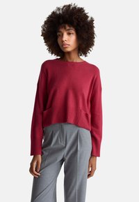 Czerwony sweter dziergany na drutach z prążkowanym dołem i długimi rękawami, zestawiony z jasnoszarymi plisowanymi spodniami. Miękka tekstura i luźny krój.