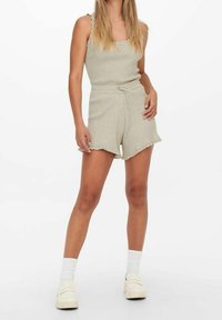 Mono corto de punto beige con costillas, tirantes finos ajustables, cintura con cordón y dobladillo deshilachado, combinado con calcetines blancos y zuecos casuales.