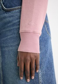 Haut rose clair à manches longues avec des poignets côtelés, orné du logo brodé "Lee". Main reposant sur un denim bleu profond, mettant en valeur la texture du tissu.