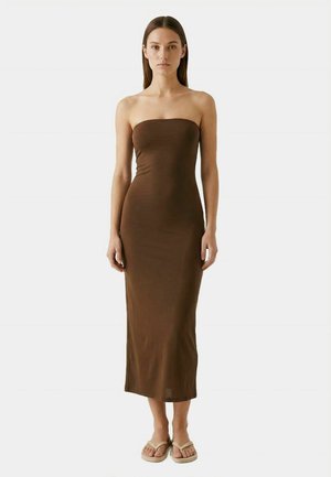 Žena s rovnými hnedými vlasmi, oblečená v hnedých midi šatách bez ramienok a béžových sandáloch, stojaca pred jednoduchým svetlým pozadím.