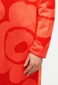 Χέρι που ακουμπά σε κόκκινο φόρεμα με μεγάλα αφηρημένα λουλουδένια σχέδια σε αποχρώσεις φωτεινού κόκκινου και πορτοκαλί.