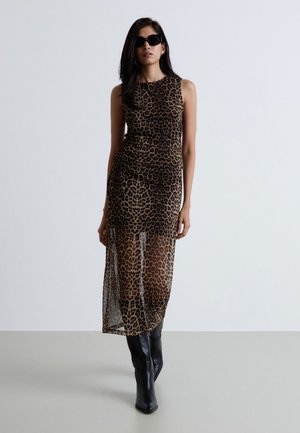 Donna con abito senza maniche a stampa leopardata con parte inferiore trasparente, stivali neri e occhiali da sole neri, in piedi su uno sfondo semplice.