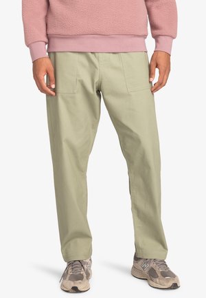 Personne portant un pantalon droit vert clair avec des poches plaquées à l'avant, un sweat-shirt polaire rose et des baskets grises, debout devant un fond blanc.