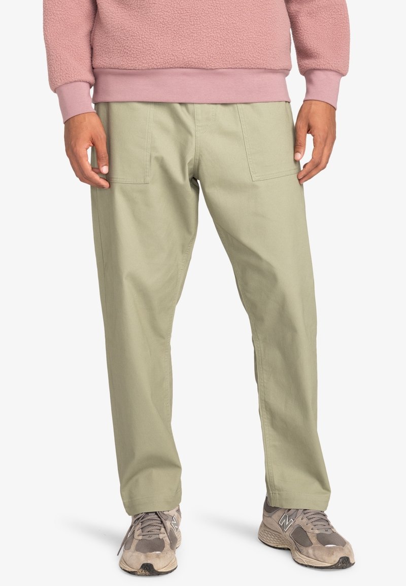 Personne portant un pantalon droit vert clair avec des poches plaquées à l'avant, un sweat-shirt polaire rose et des baskets grises, debout devant un fond blanc.