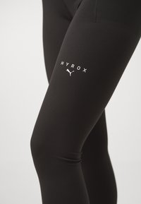 Leggings atletici neri con una consistenza liscia, con un logo bianco "HYROX" e un emblema Puma sulla coscia.