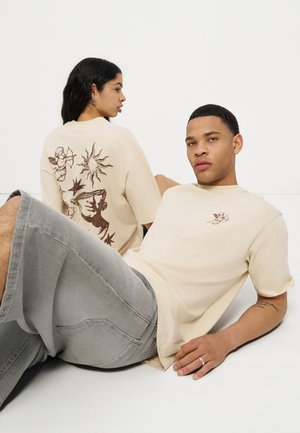 Due modelle che indossano t-shirt grafiche color crema, una seduta di fronte e l'altra con le spalle rivolte mostrando disegni di sole, stelle e angeli.