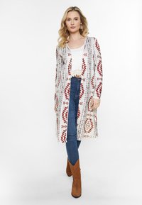 Langer, leichter Cardigan mit roten und braunen geometrischen Mustern auf hellem Hintergrund, kombiniert mit einem weißen Oberteil und dunkelblauen Jeans.