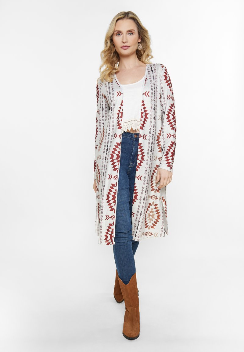 Langer, leichter Cardigan mit roten und braunen geometrischen Mustern auf hellem Hintergrund, kombiniert mit einem weißen Oberteil und dunkelblauen Jeans.